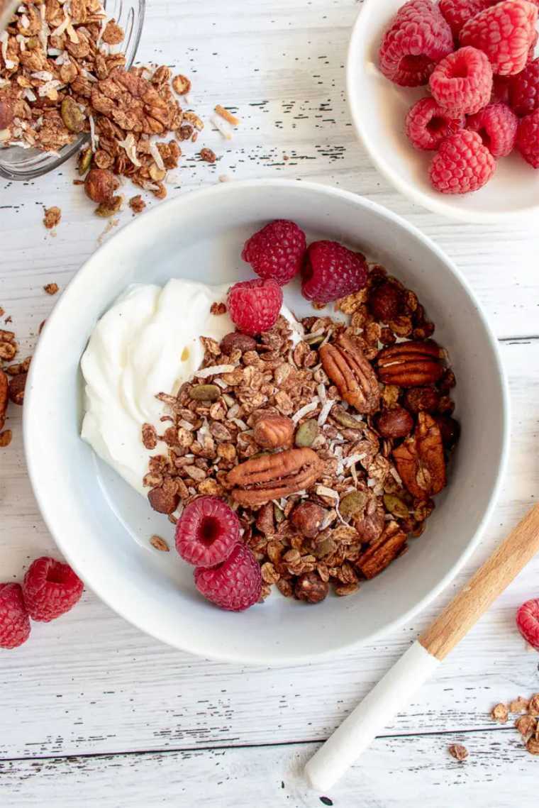 white bowl on a table filled with vegan chocolate granola, coconut yogurt and raspberries