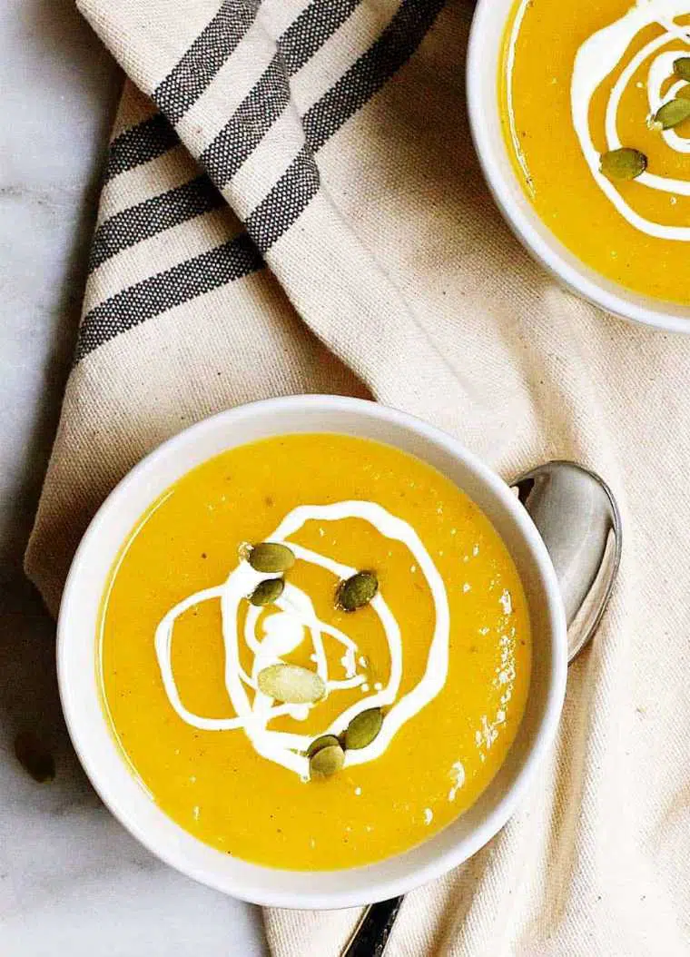 linen towel with two white bowls full of orange vegan butternut squash soup topped with seeds