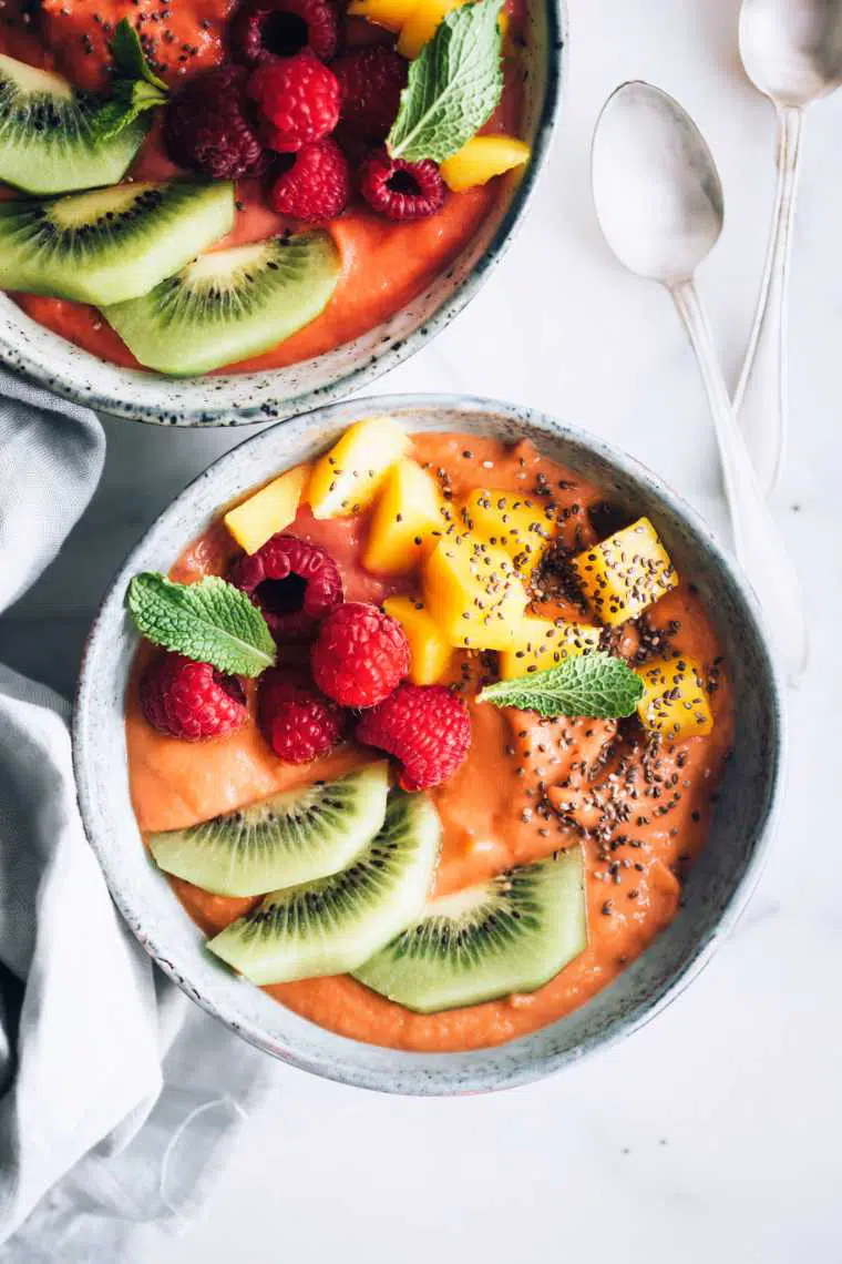 white table with two bowls that are filled with orange sweet potato smoothie, kiwi, mango and raspberries