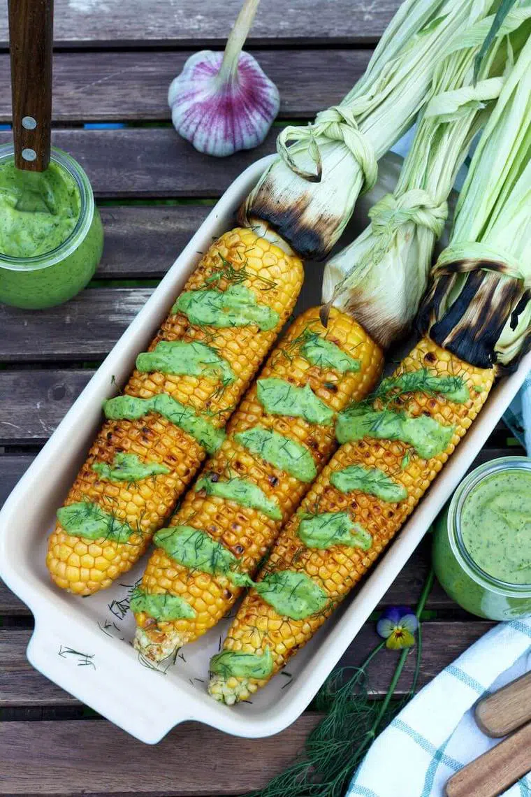white baking dish on a table filled with three grilled corn on the cob that are drizzled with some avocado dill dressing