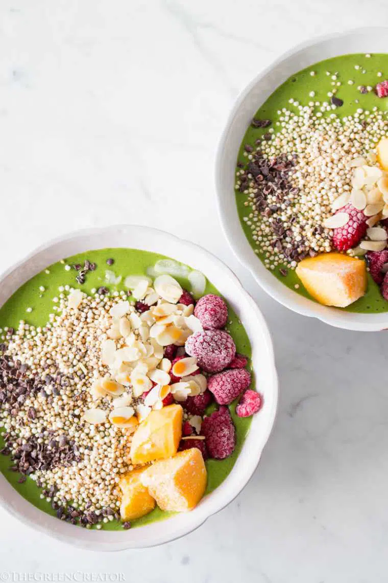 white table with two bowls filled with green smoothie and topped with fruit, nuts and quinoa