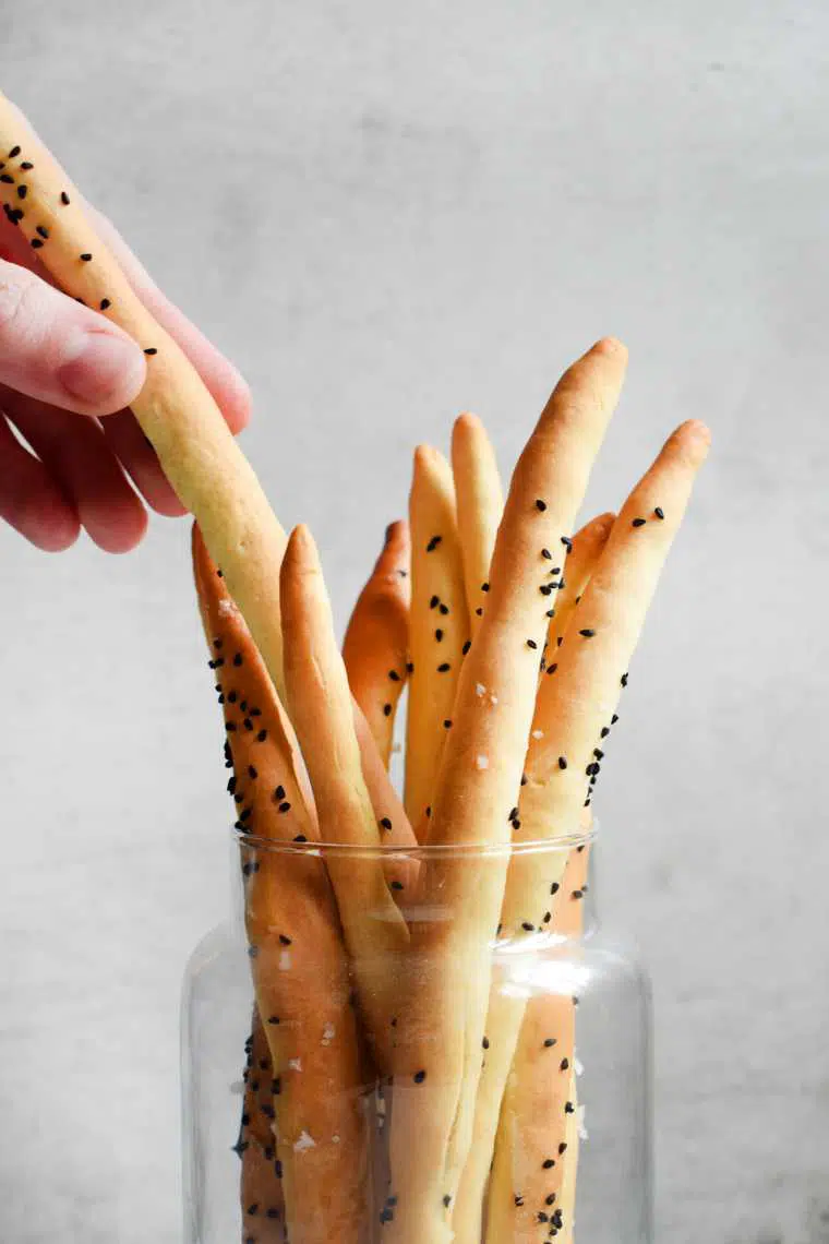 glass with almost a dozen homemade vegan breadsticks one of which is taken by a hand