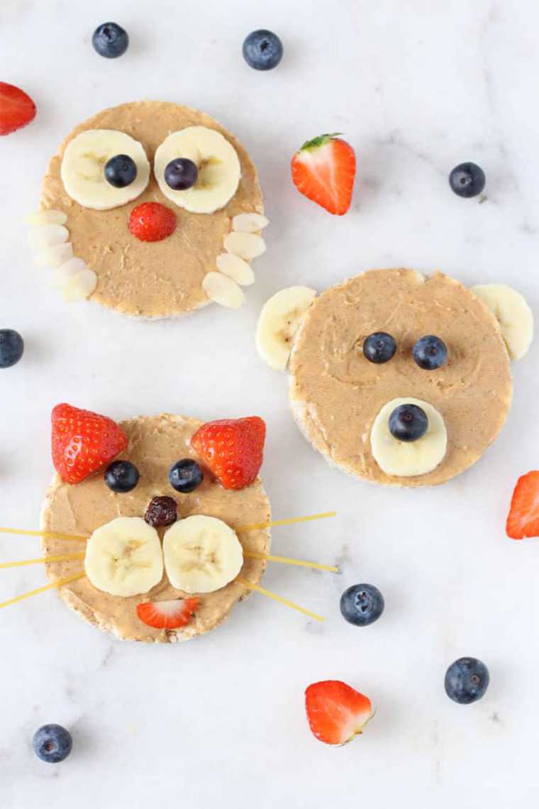 white table with three rice cakes dressed with peanut butter and fresh fruit to look like animals