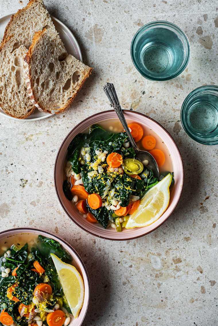 two rose-colored bowls on a table filled with kale, white bean, carrot and lemon soup for a light vegan dinner