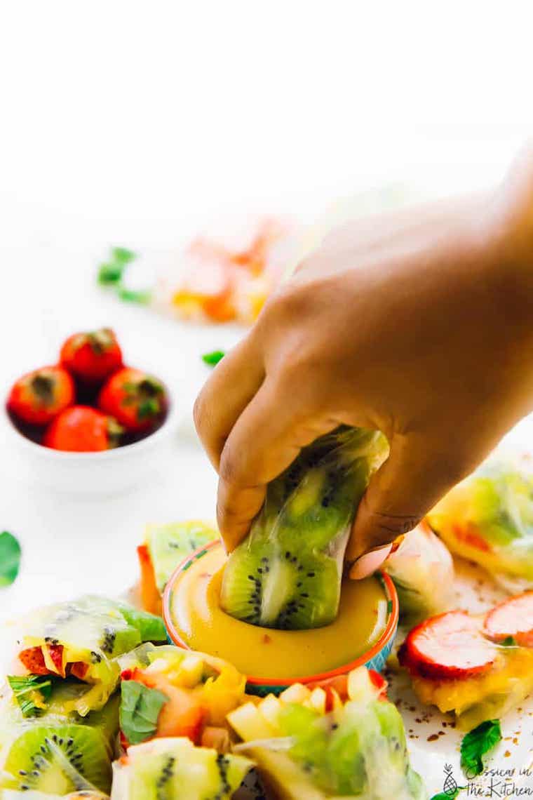 colorful fruit spring rolls wrapped in rice paper where one is taken by a hand and dipped in mango dressing