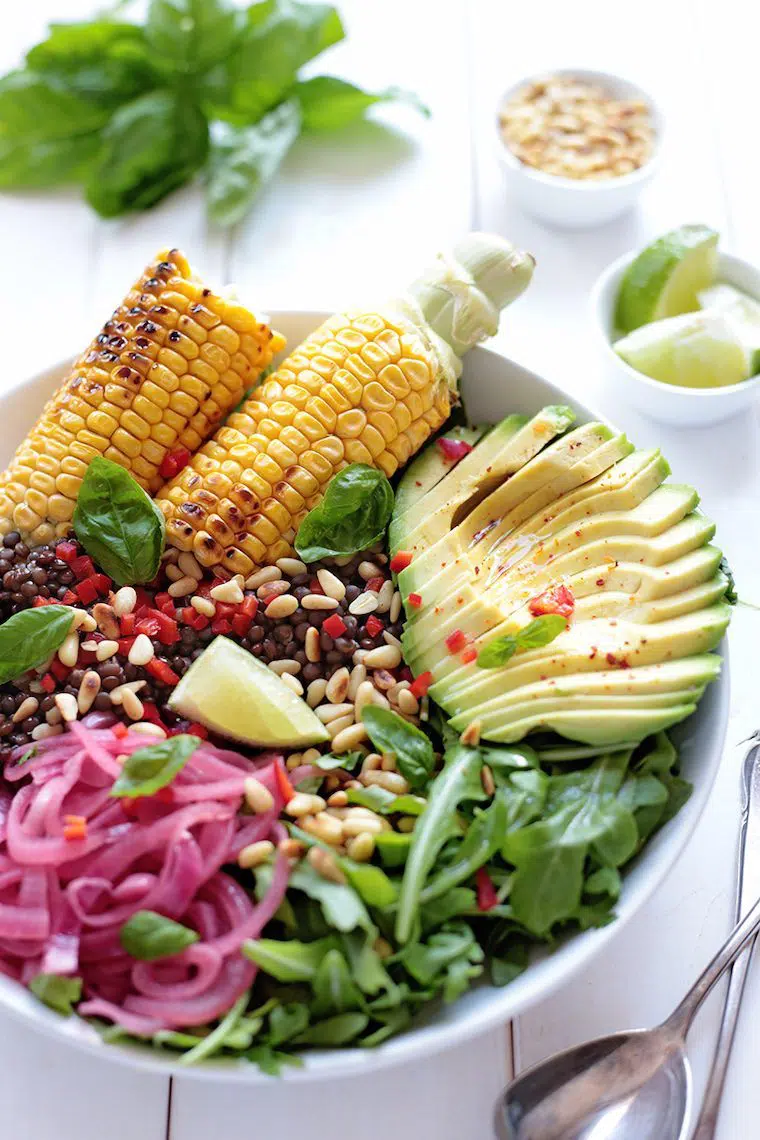 large white bowl with leafy greens, sliced avocado, pickled onion, lentils, grilled corn and pine nuts