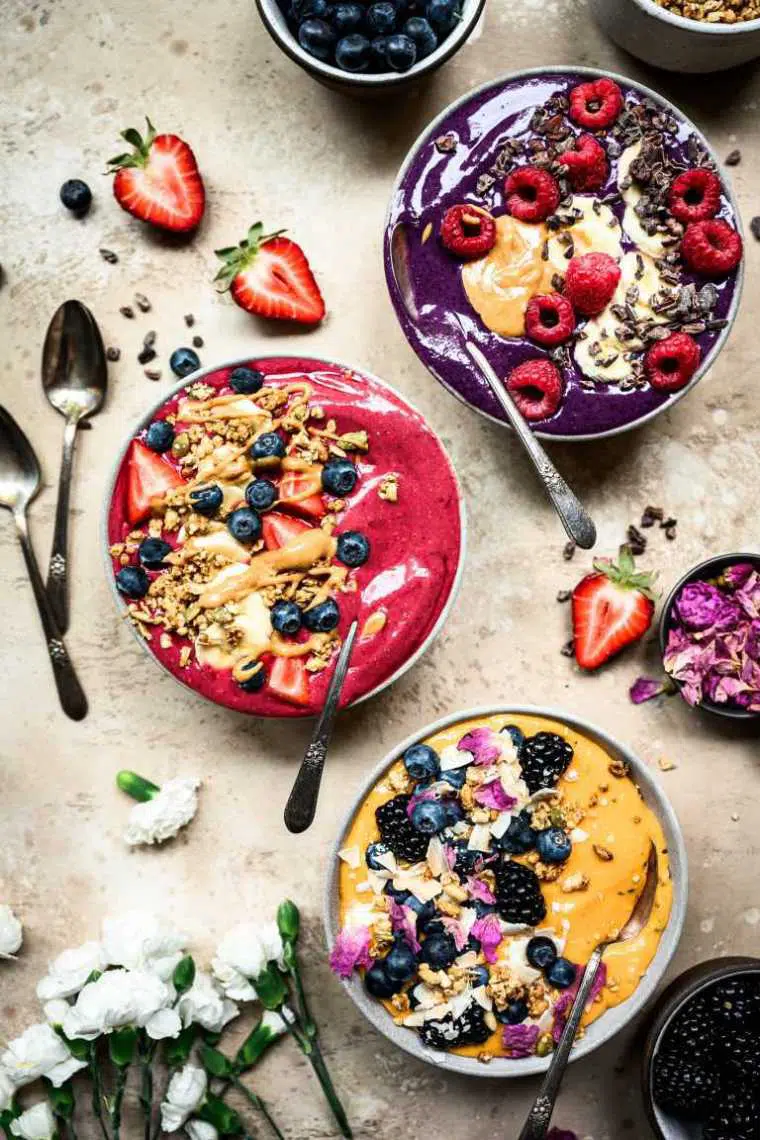 top view of table with white flowers, several spoons, berries and three colorful bowls filled with superfood smoothies and healthy vegan toppings