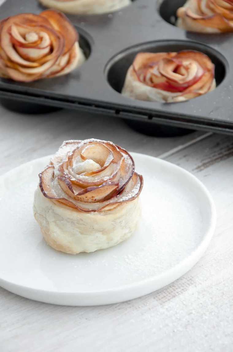 white table with one vegan apple rose made from puff pastry next to a baking pan with more roses