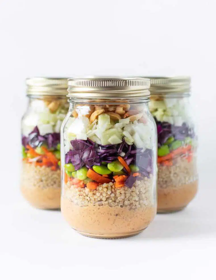 three mason jars with layered peanut crunch salad containing quinoa, edamame and red cabbage