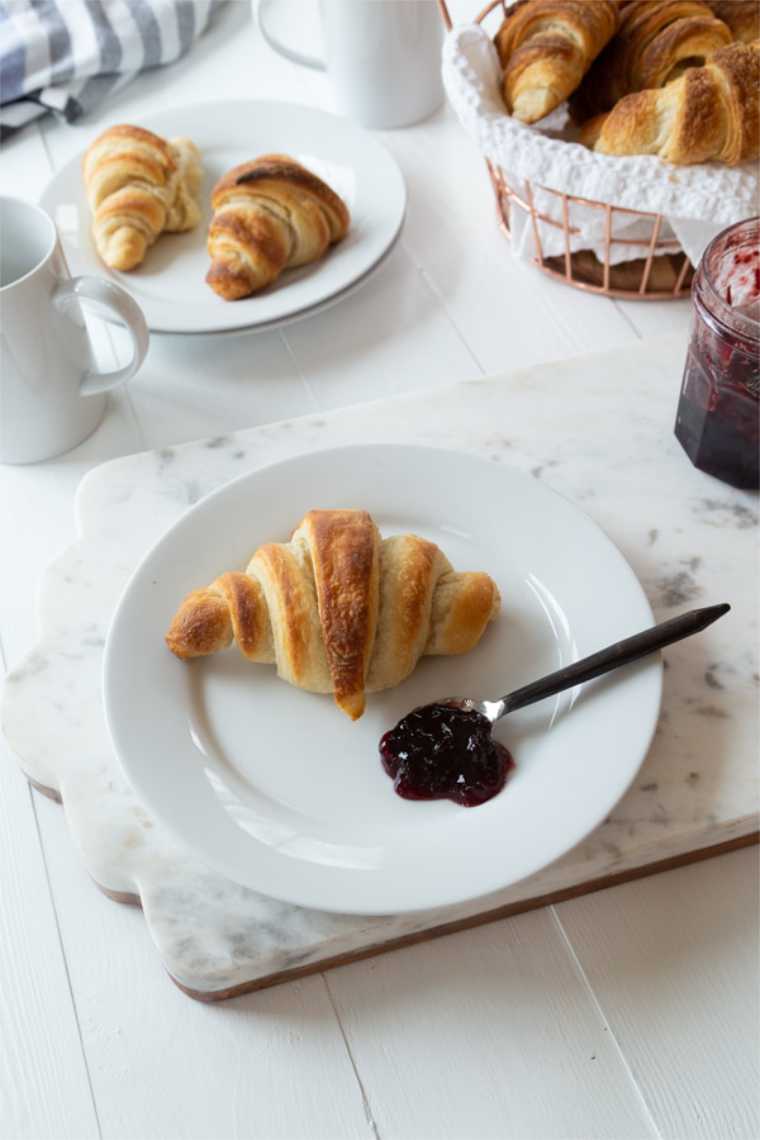white table with cups, jam, cutting board and two white plates featuring homemade vegan french croissants