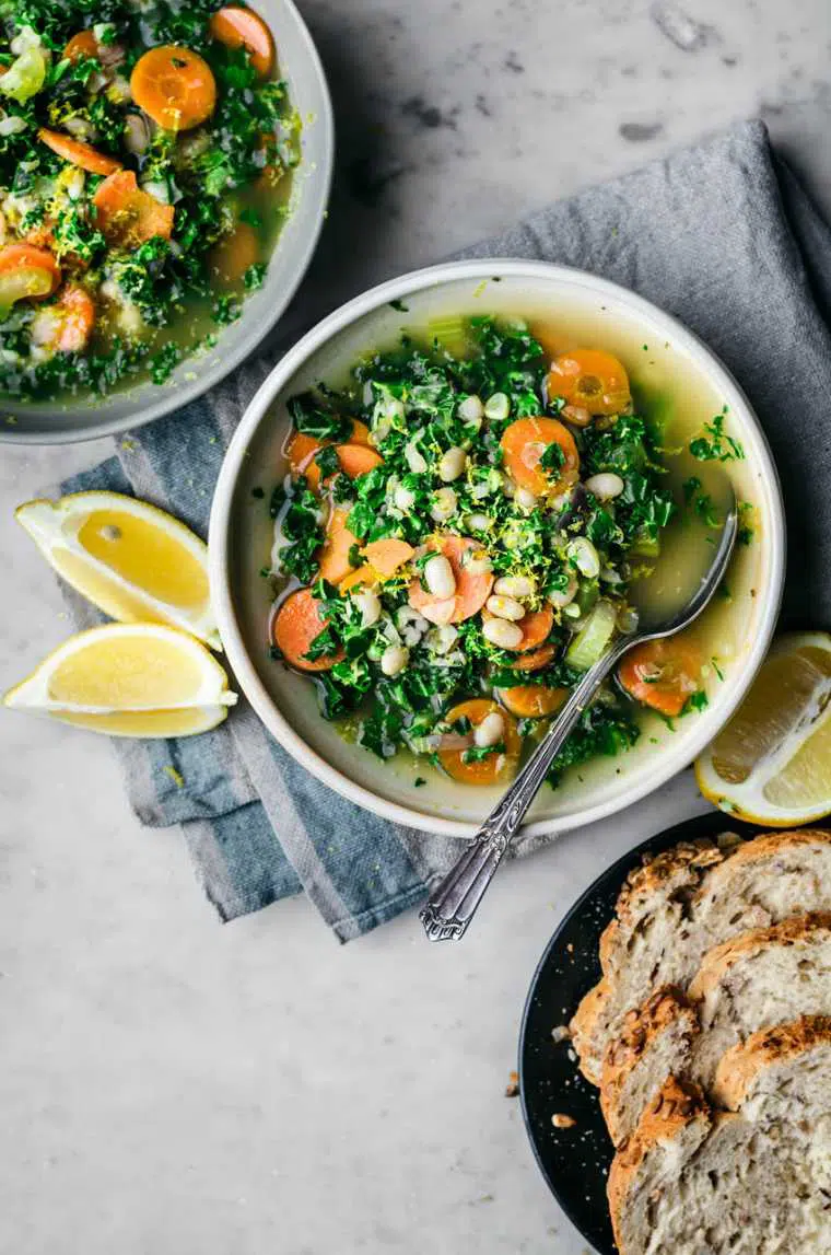 two bowls with plant-based White Bean, Lemon and Kale Soup and a spoon next to sliced bread