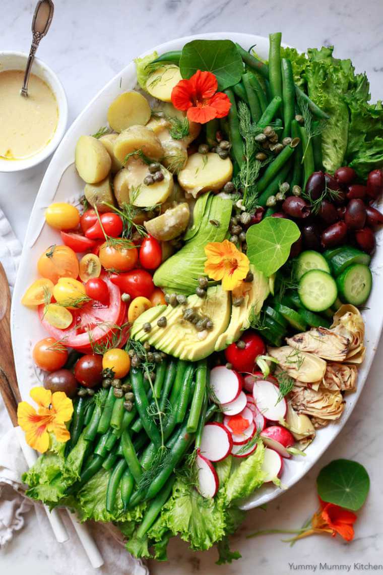 large white platter with nicoise salad featuring potatoes, green beans, avocado, olives, tomatoes and radishes
