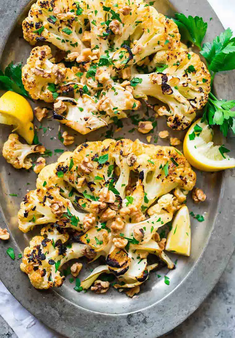 silver plate with two crispy grilled vegan cauliflower steaks, walnuts, herbs and slices of lemon