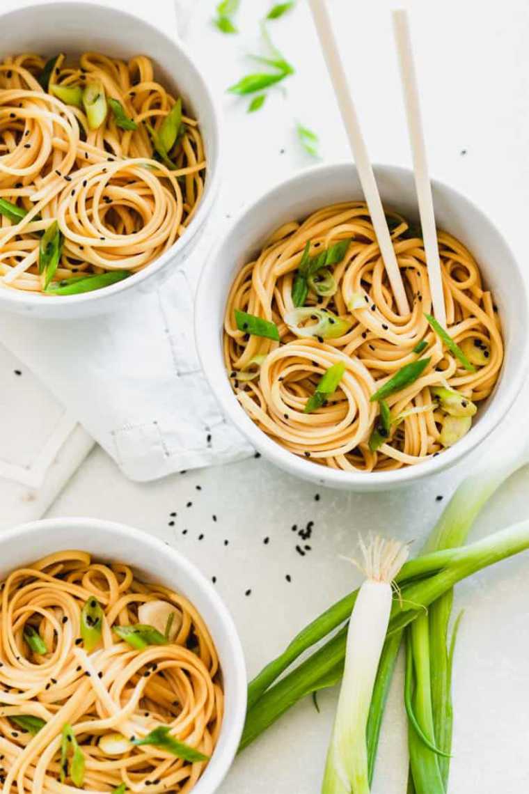 three small bowls with sesame noodles next to some green onion