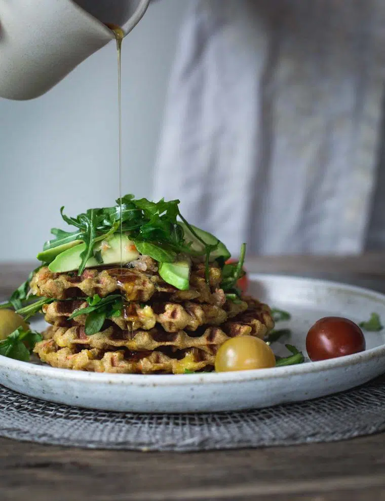 white plate of 4 mashed potato cheese waffles on top of each other with avocado, tomatoes, rocket and maple syrup being poured over everything