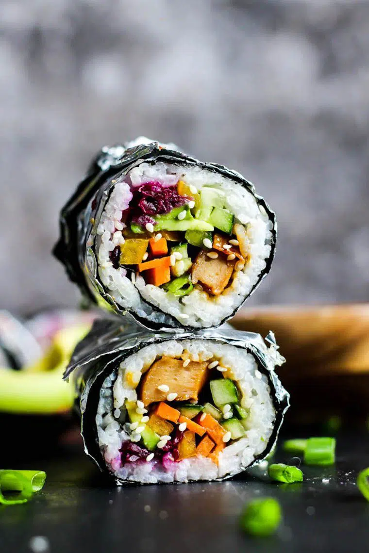 two sushi burritos on top of each other containing tofu, carrot, avocado, rice and other veggies
