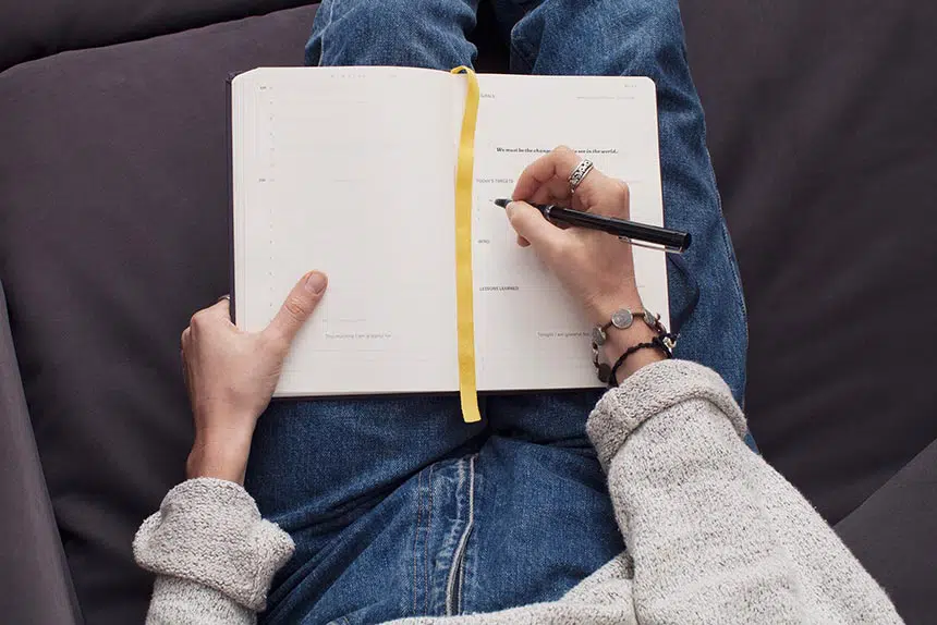 woman with cozy beige sweater and jeans sitting on the soda and writing in her journal to create healthy habits