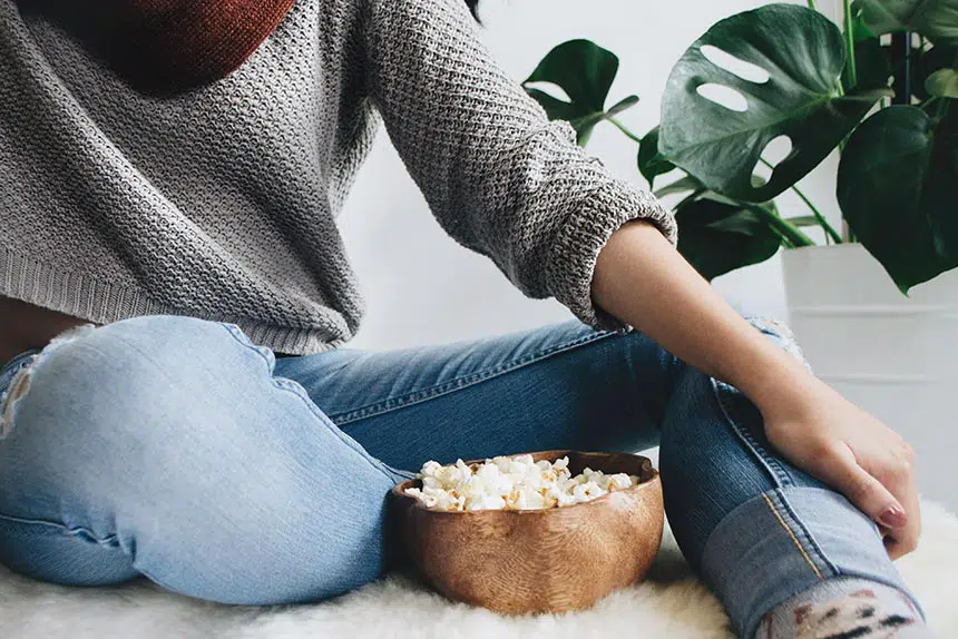 woman in tight jeans and grey knitted sweater sitting next to a wooden bowl of popcorn