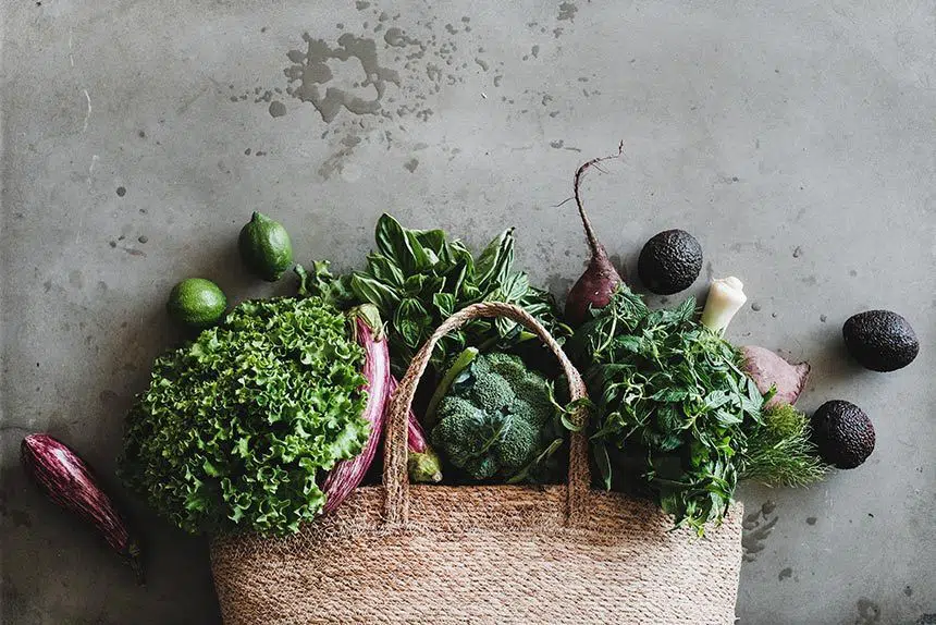 textile grocery bad laying on a wooden surface with fresh produce such as leafy greens, herbs, avocado, broccoli and aubergine