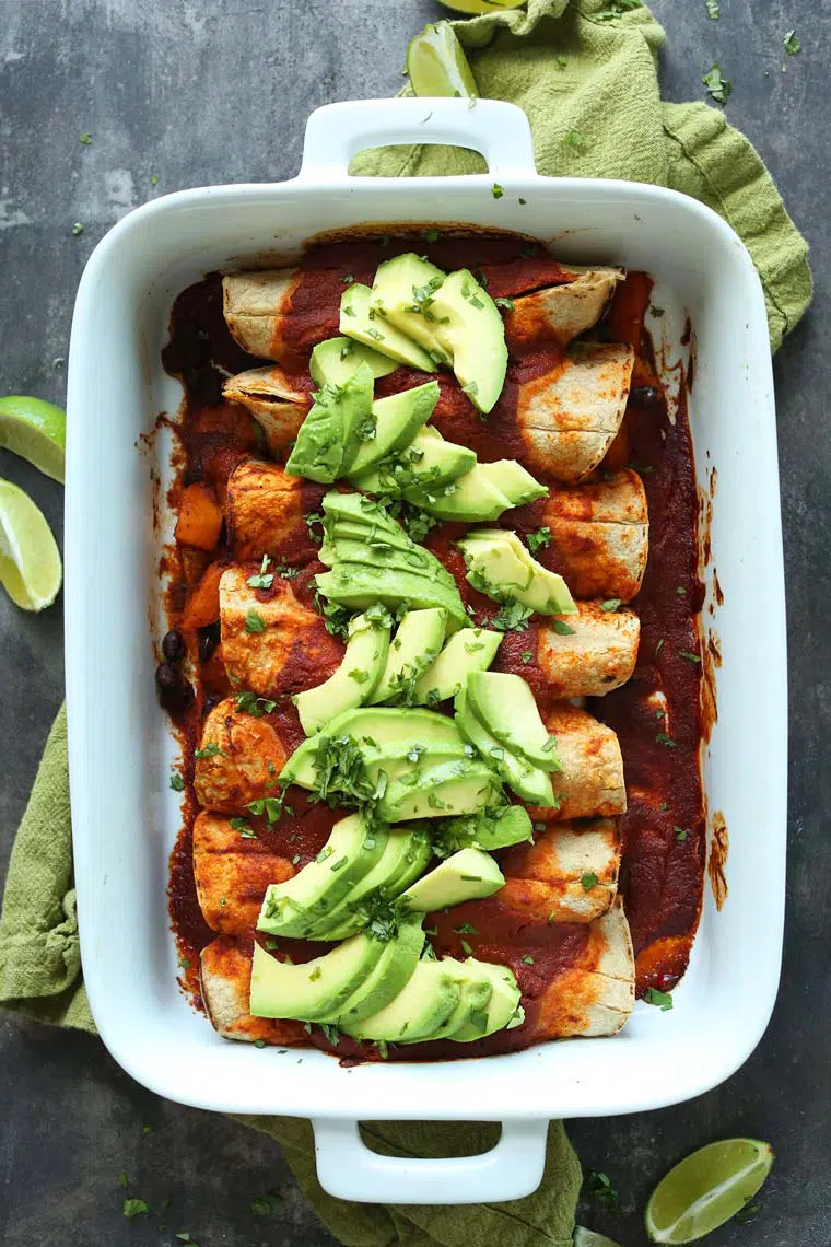 white baking dish on a green towel with homemade vegan butternut enchiladas topped with avocado