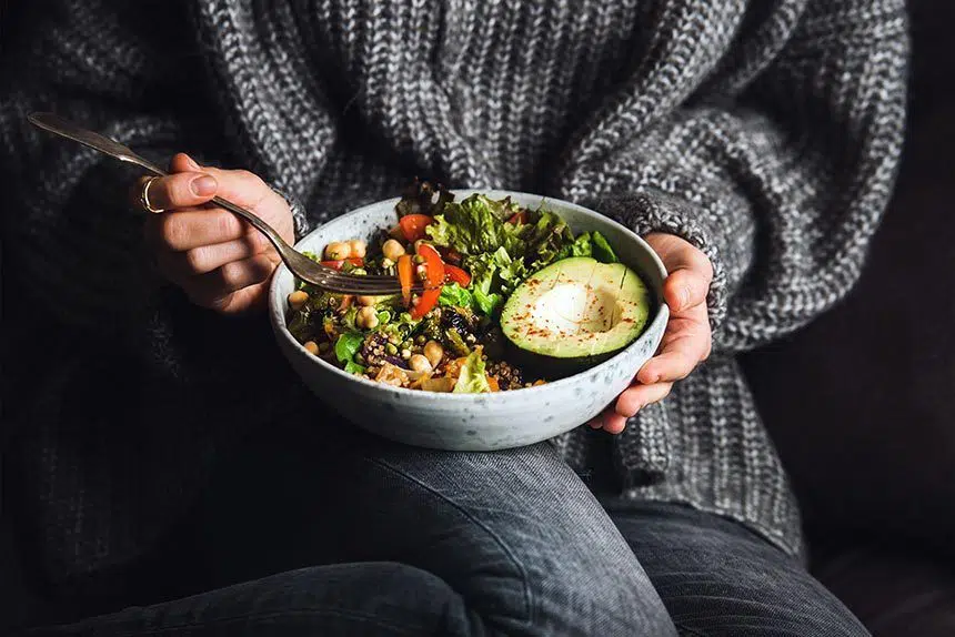 woman wearing jeans and a cozy knitted grey sweater having a ceramic bowl with greens, bell pepper, grains, beans and avocado on her lap