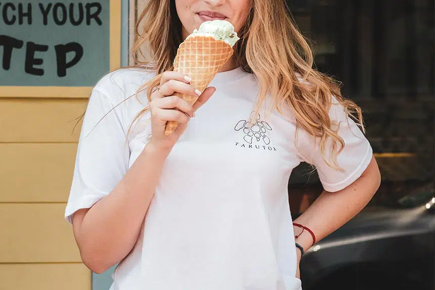 blonde woman with white tshirt standing in front of a yellow wall and smiling while eating ice cream out of a cone