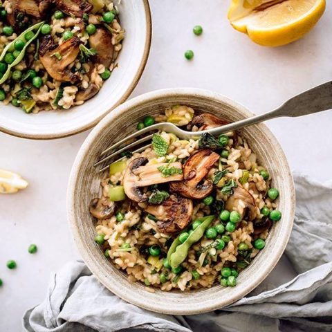 bowl of vegan risotto with mushroom, peas and a fork standing next to a linen on a marble surface