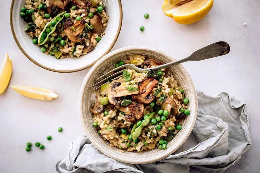 bowl of vegan risotto with mushroom, peas and a fork standing next to a linen on a marble surface