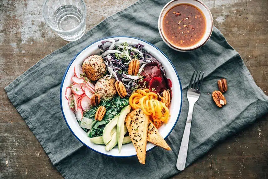 white bowl with colorful plant based food such as tofu, radishes, avocado, greens, nuts and cabbage standing on a linen next to a brown sauce
