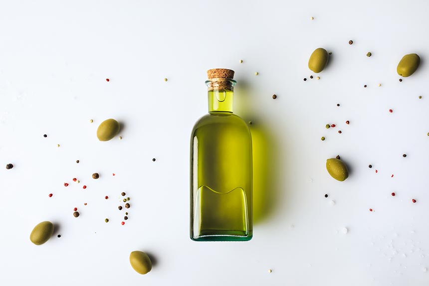 glass bottle of olive oil lying on a white surface next to some olives and pepper