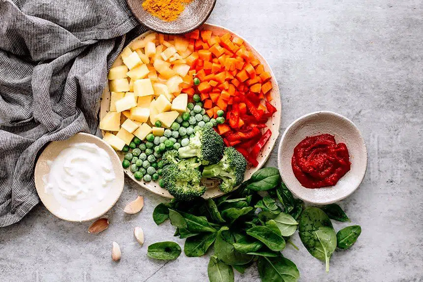 plate with potato, carrot, bell pepper, broccoli and peas standing next to small bowls of soy yogurt, spices, tomato paste and fresh spinach