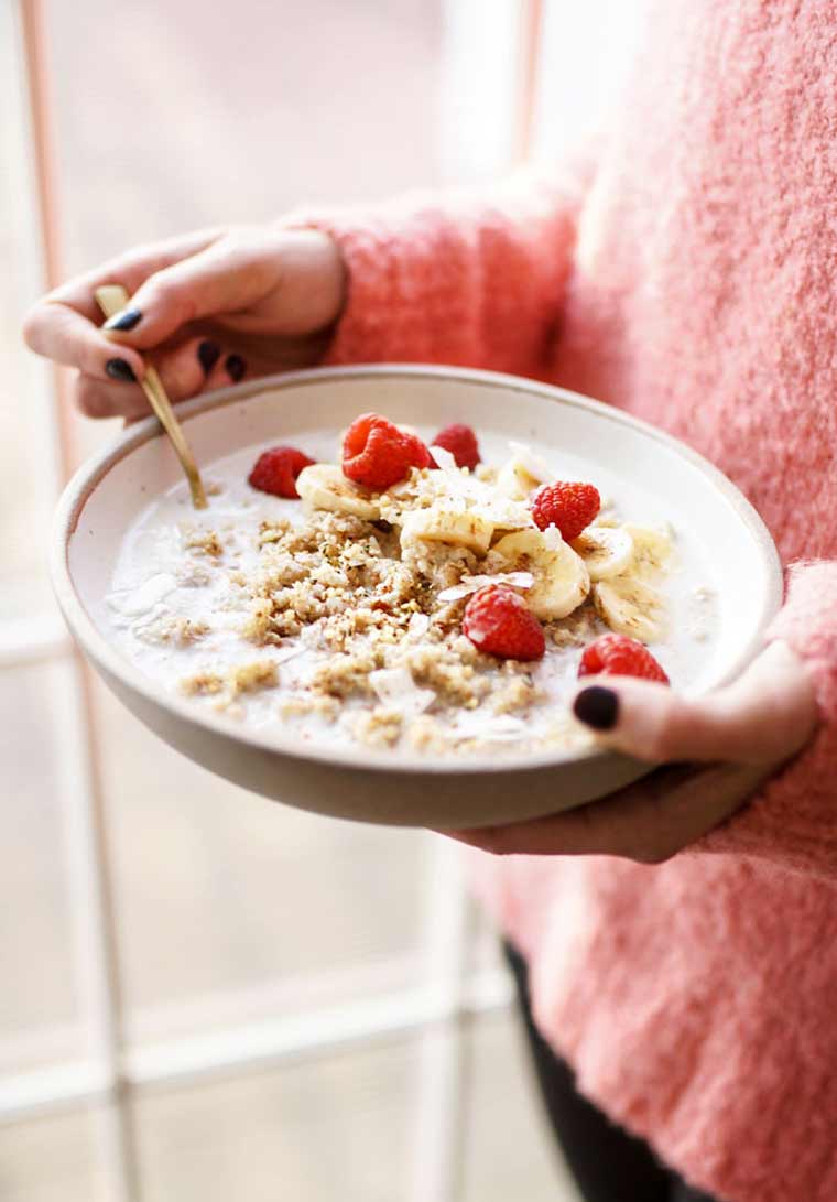 woman in pink sweater standing by the window and holding a bowl of cooked quinoa and fruit