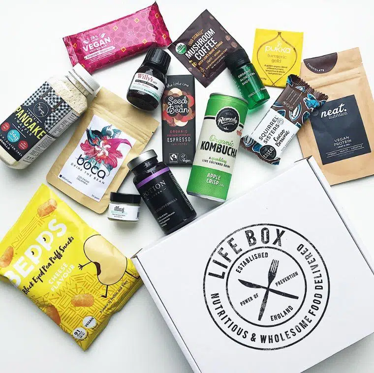 Overhead view of white box with Lifebox printed on it and a variety of vegan snacks and drinks arranged around it such as protein pancake mix, black eyed pea puffs and mushroom coffee
