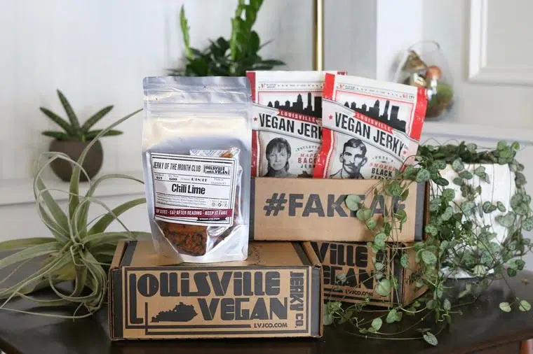 Three boxes of vegan jerky on a dark surface surrounded by plants