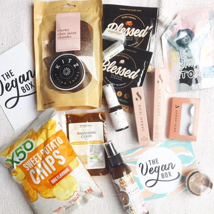 Overhead view of the contents of The Vegan Box arranged on a white fabric: sweet potato chips, protein powder, choc mint chunks and several skin care products