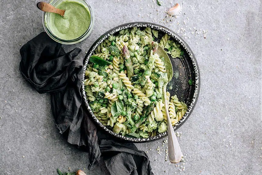 dark bowl of vegan pesto pasta with steamed broccoli and asparagus next to some oil free vegan pesto