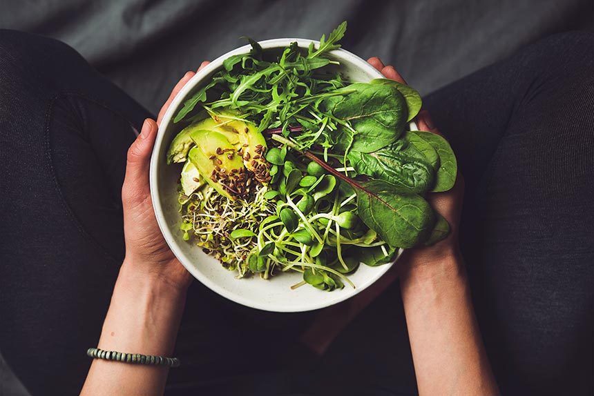 woman in black leggings sitting cross-legged and holding a white bowl of leafy greens, sprouts and avocado in her hands