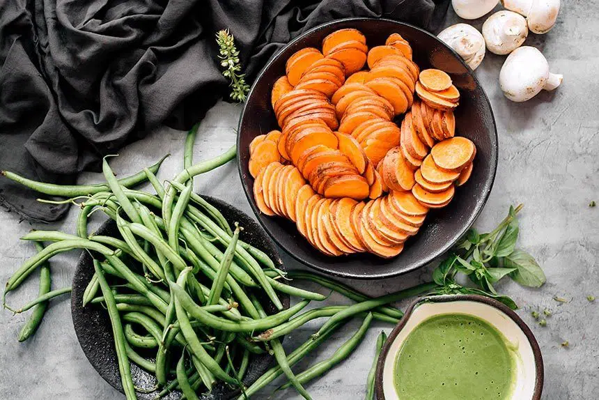 two black bowls filled with green beans and sweet potatoes standing on a stone surface next to some mushrooms and green sauce