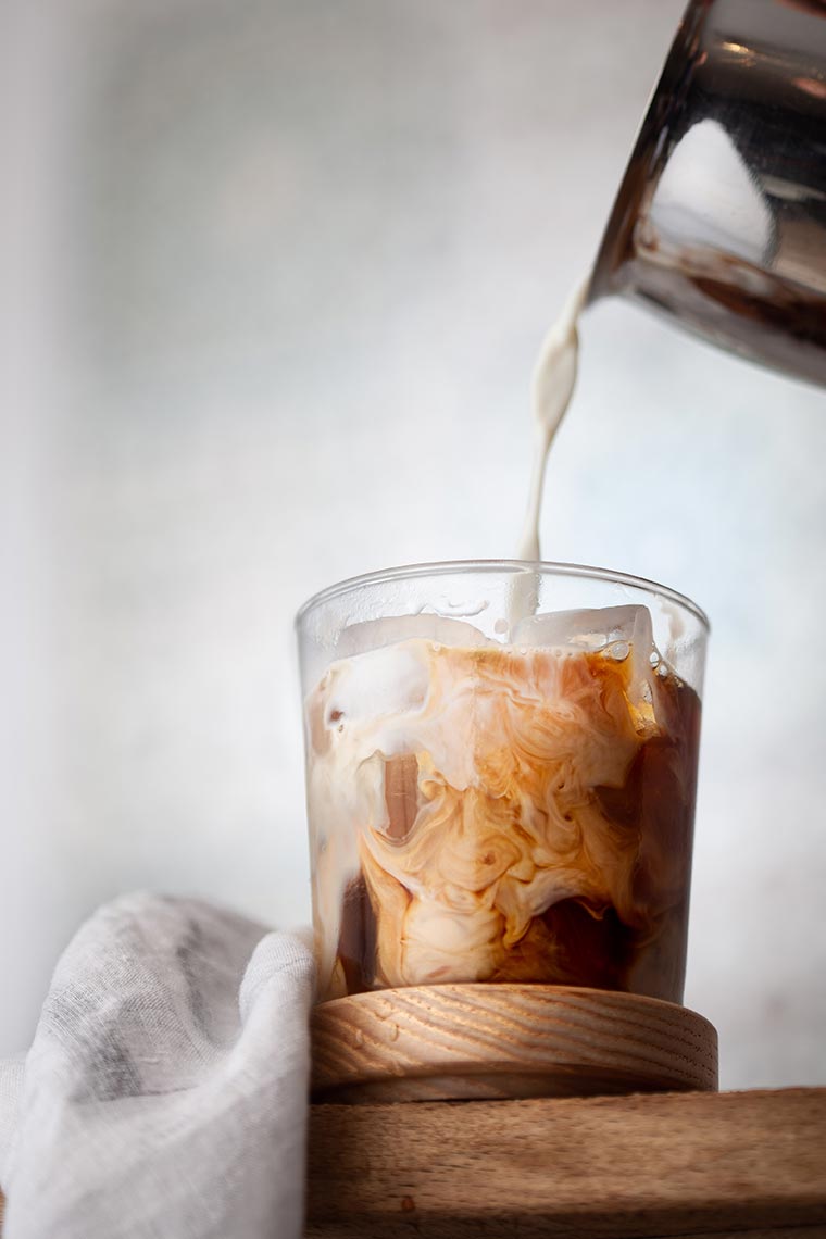 pouring soy milk in a glass of cold coffee with ice cubes standing on a wooden table