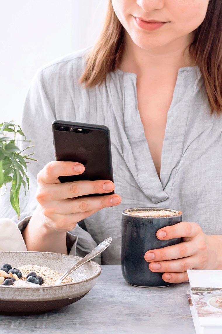 woman in grey shirt sitting on a table and looking at her phone while drinking almond coffee
