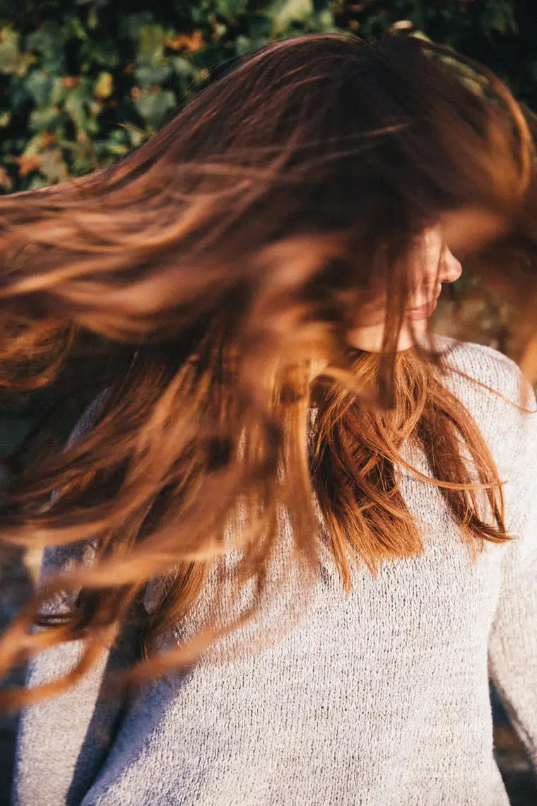 long brown-red haired woman wearing a grey sweater shaking her head and long hair in the sun