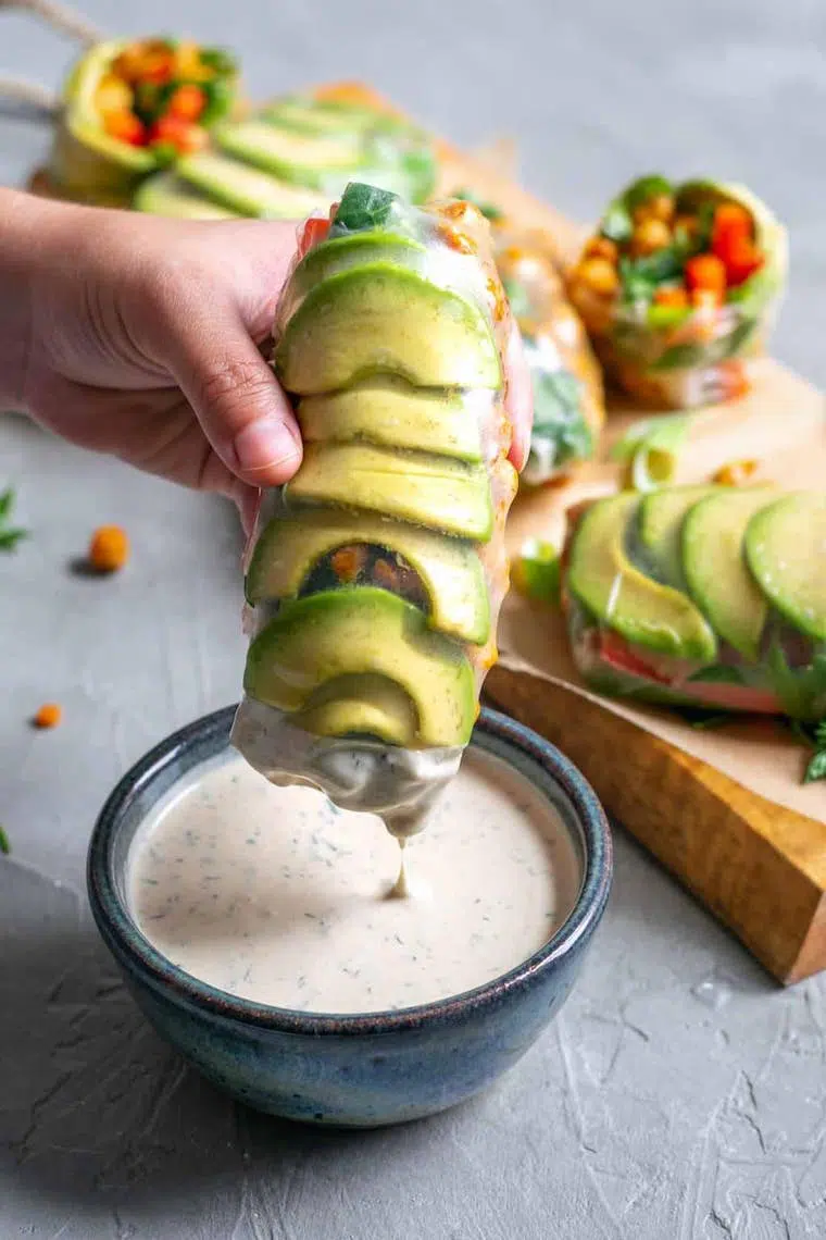 hand holding a vegan rice paper wrap with lots of avocado and dipping it in a creamy sauce