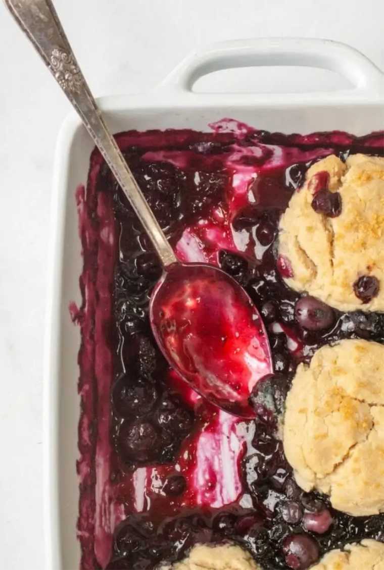 close up of a white baking dish with vegan blueberry cobbler and a spoon