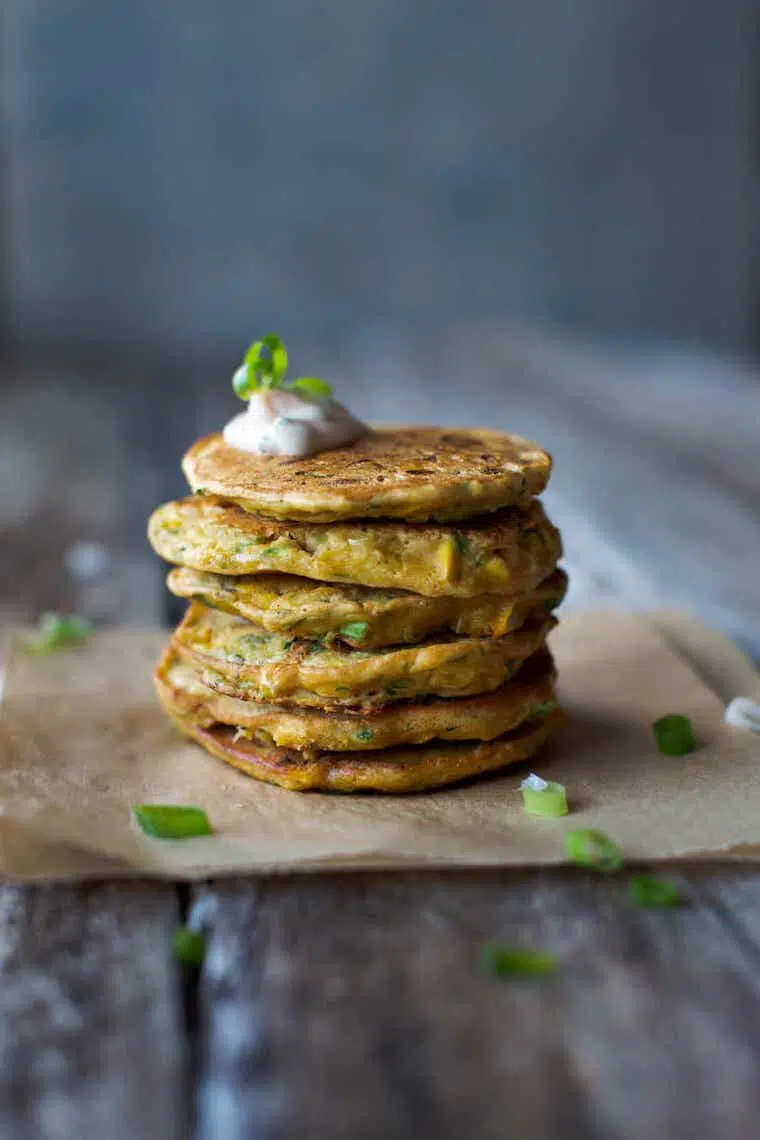 piece of parchment paper on a wooden table with six vegan corn and zucchini fritters stacked on top of each other