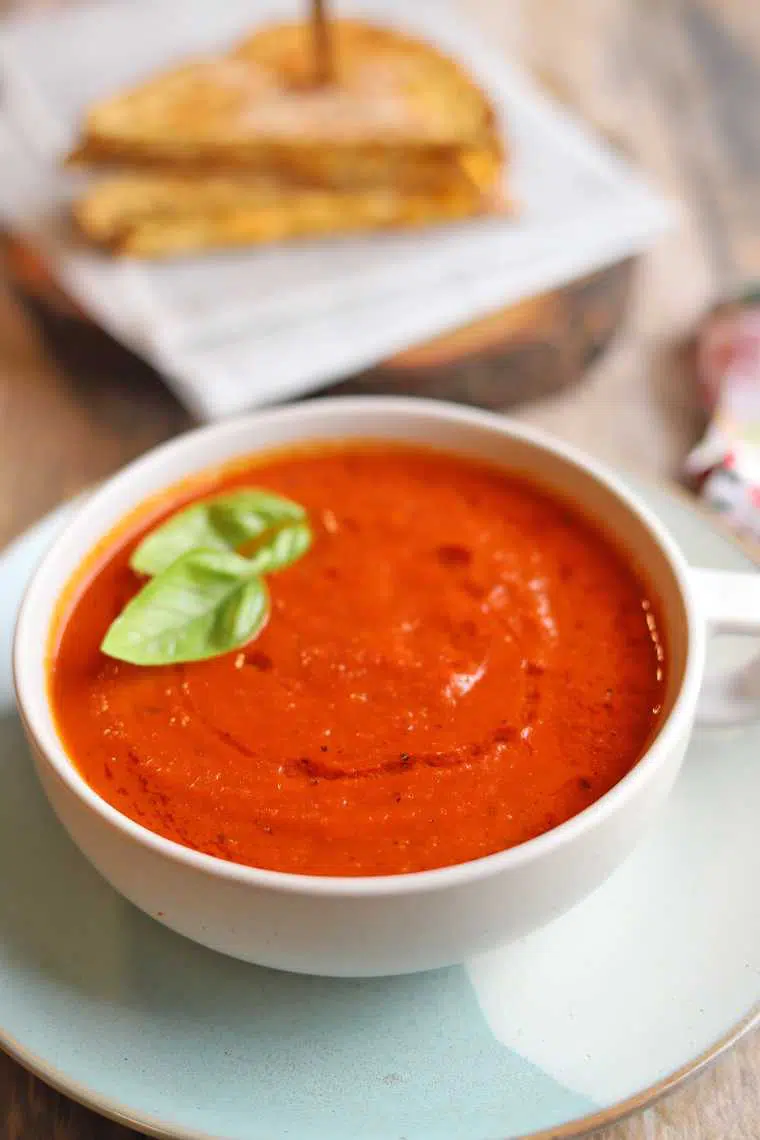 white bowl of classic vegan tomato soup with some basil next to a vegan sandwich