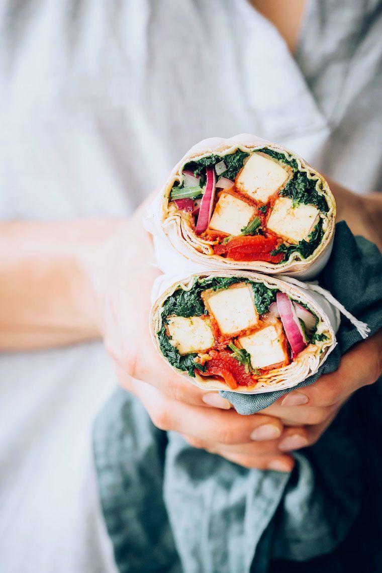 woman in linen shirt holding a blue towel as well as two tofu and spinach stuffed vegan wrap