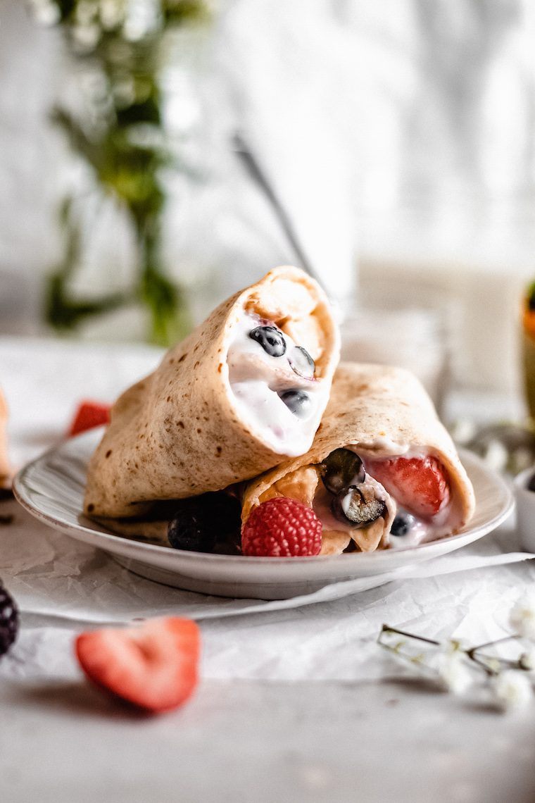 breakfast table with a white plate on which sweet soy yogurt and berry stuffed vegan crepes lie