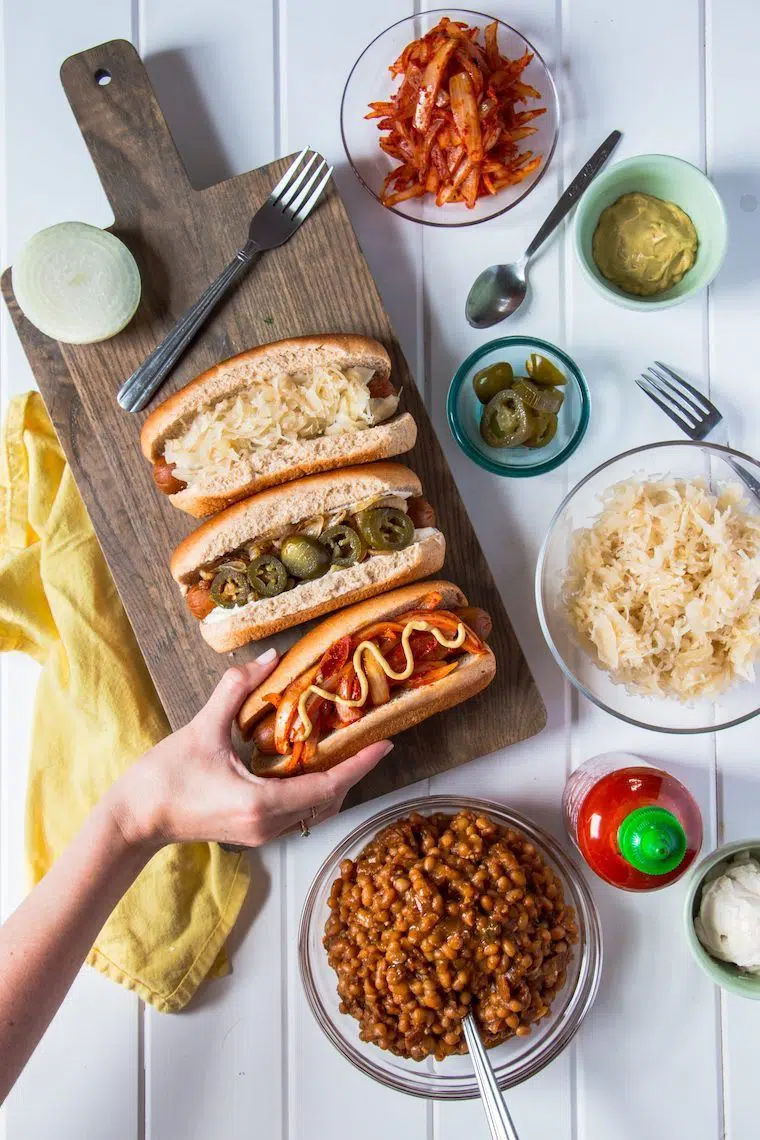 white table with a wooden chopping board and three different kinds of vegan hot dogs next to bowls with ingredients to make them