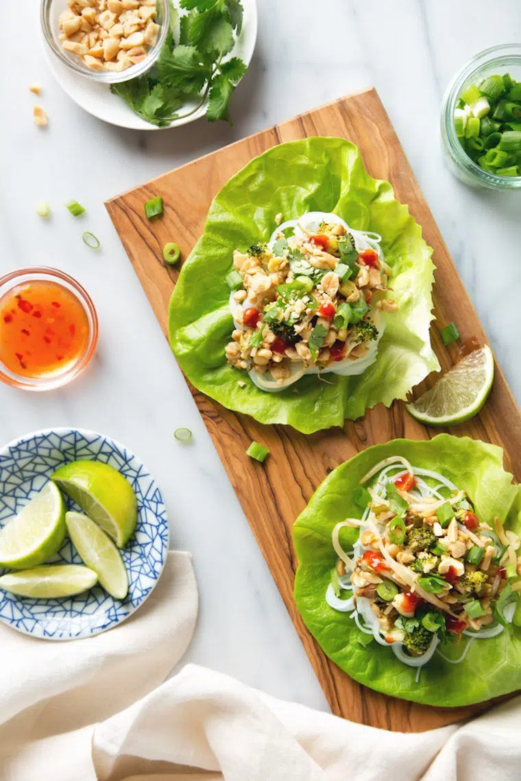 white table with wooden chopping board with open lettuce wraps that are filled with glass noodles and Thai flavored veggies