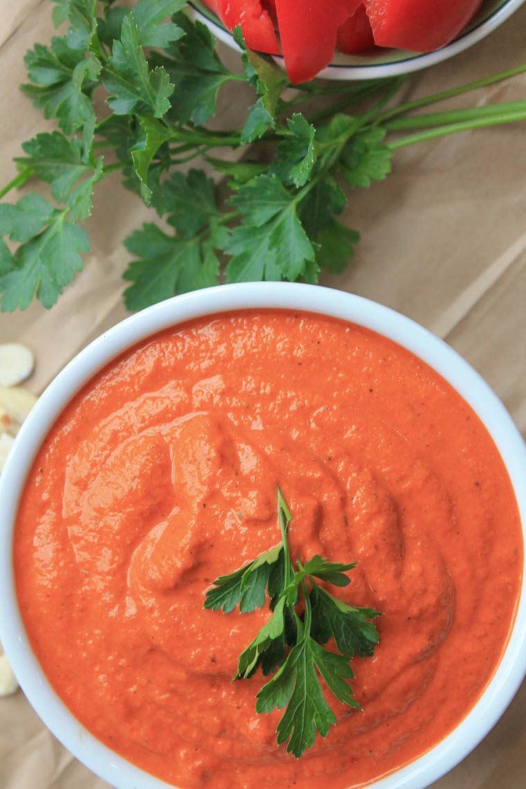 table with some bell pepper, fresh herbs and a white bowl filled with red vegan romesco sauce